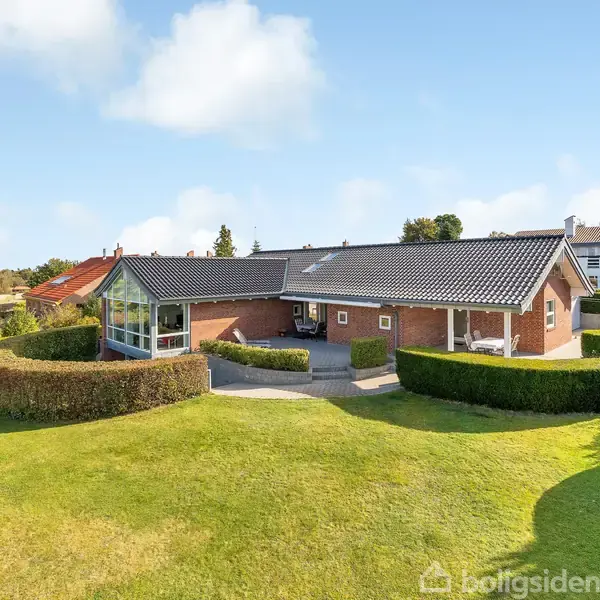 En rød murstensvilla med glasudestue er omgivet af en velplejet have med klippede hække og græsplæne. Huset ligger i et villakvarter med flere omkringliggende hjem.