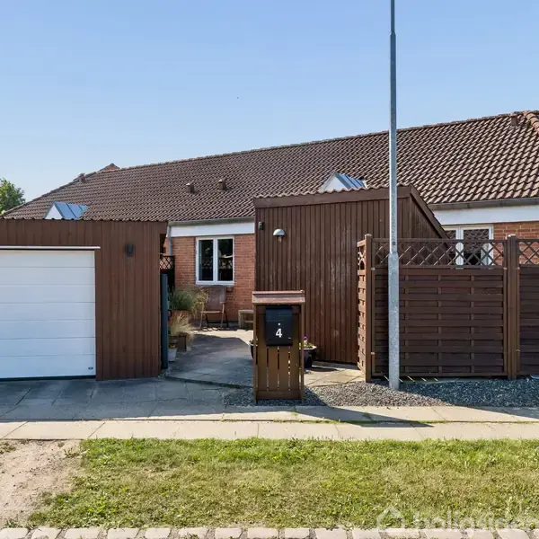 Hus med rødt tag og træfacader står bag en forhave med en indkørsel og garage. Der er et skilt med tallet "4" ved en postkasse foran indgangen.