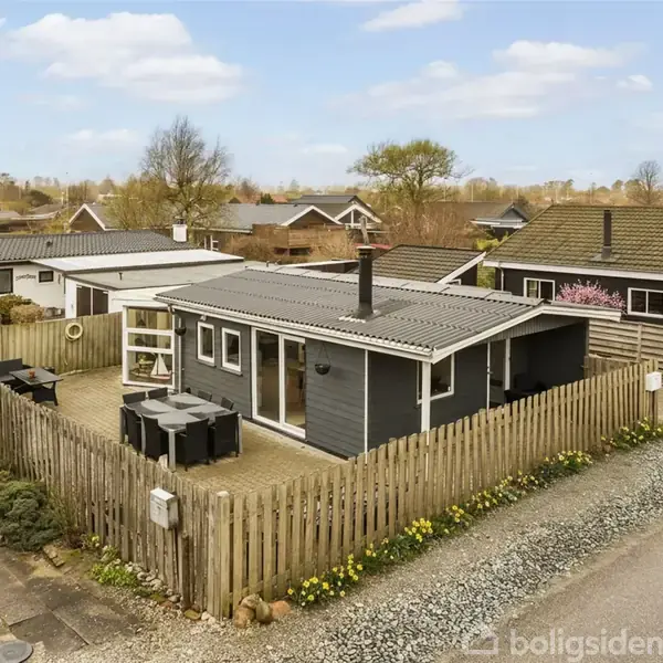 Et sortmalet sommerhus står stille omgivet af en lav træhegn. På terrassen står havemøbler. Hytten ligger i et villakvarter med flere ensartede bygninger og træer i baggrunden.