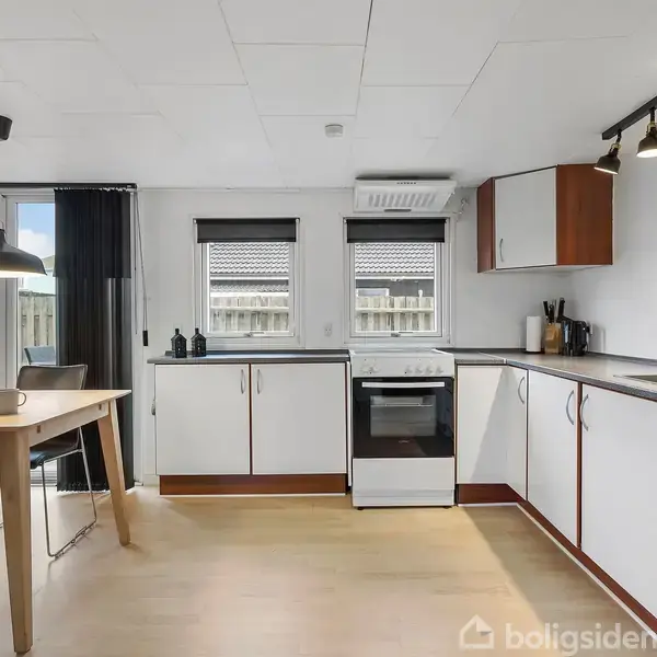 Køkken med hvide skabe og trædetaljer, komfur og ovn som centrale elementer, omgivet af spisebord og stole. Store vinduer lader lys ind, og der er udsigt til en terrasse.