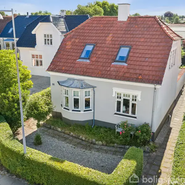 Hvidt hus med rødt tag, der har skråvinduer. Huset ligger i en haveramme med hække og grusbelagt forhave. Omgivet af andre lignende boliger i et villakvarter.