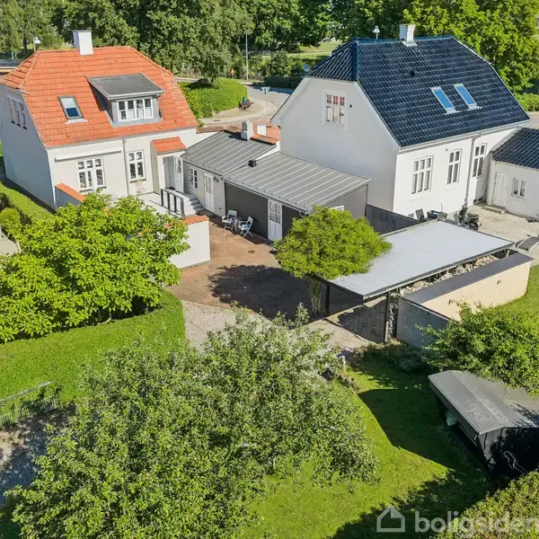 Huse med røde og sorte tage står side om side i et kvarter, omringet af grønne træer og græsplæner. En enkelt carport og have kan ses i forgrunden.