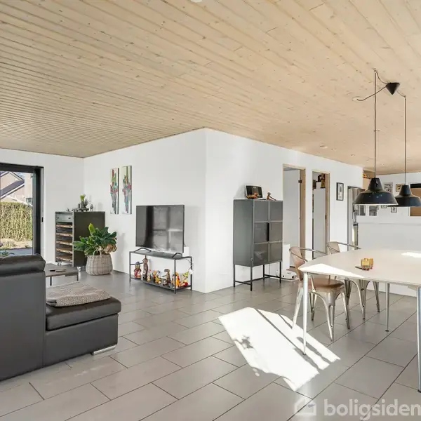 En stue med sort lædersofa og tv på et stativ. Et spisebord med stole står i nærheden. Lyse fliser og træloft præger rummet med store vinduer og åbent køkken.