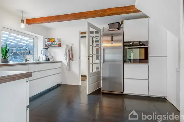 Køkken med hvide skabe og stål-køleskab, glasdør åben mod et anden rum. En træbjælke i loftet og store fliser på gulvet. Lys fra stort vindue ved køkkenbord.