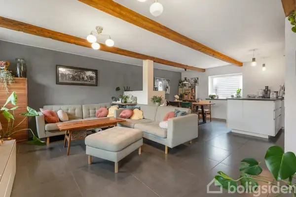 Lys stue med en stor sofa og sofabord. I baggrunden er der et moderne køkken og spiseplads. Grønne planter pryder rummet, og et maleri hænger på væggen.