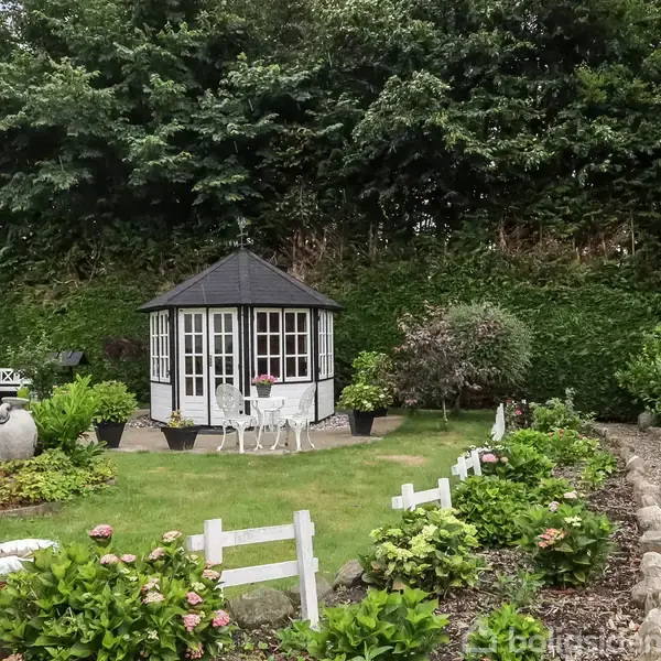 Et lille lysthus med sort tag står i midten af en velholdt have. Stier med småsten og blomsterbede omgiver huset. Tykke hække udgør baggrunden.