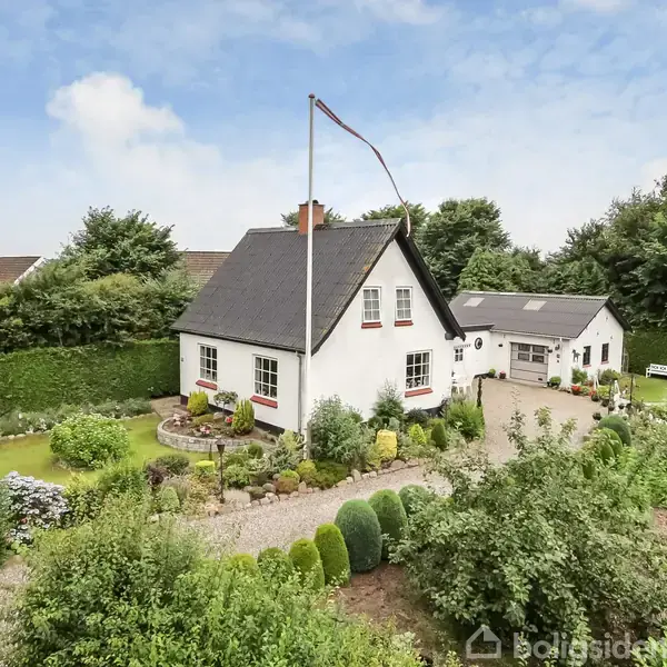 Et hvidt hus med sort tag står i en velplejet have med buske og blomsterbede. Et flagstang uden flag ses foran huset i en grøn, frodig omgivelser.