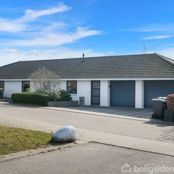 En én-etagers villa med hvid facade og gråt tag står i et boligområde. Der er en garageport og en forhave med buske. Himlen er blå med lette skyer.