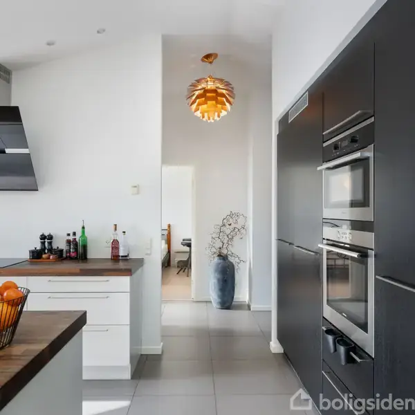 En moderne køkken med mørke skabe og indbyggede apparater; en frugtskål står på bordet. En lampe hænger i gangen; en vase med grene ses i baggrunden.