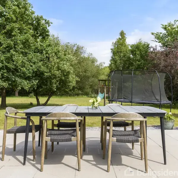 Et havebord med seks stole står på en flisebelagt terrasse. Ved siden af ses en trampolin. Omgivet af frodige grønne træer og en græsplæne.