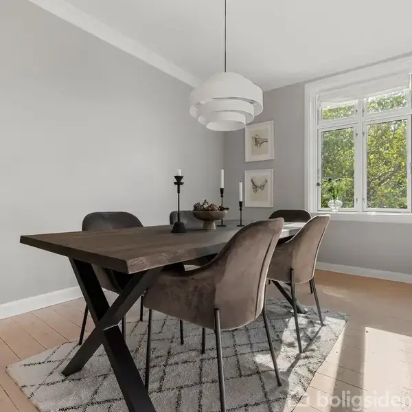 Spisestue med seks stole omkring et træbord, oplyst af en moderne pendellampe. Foran ses en rund marmorbordplade med dekorationer. Loftroset i hvidt som detaljeret design i højre hjørne.