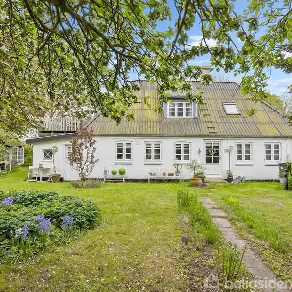 Hvidt hus med gammeldags charme står stille i en frodig have med blomsterbede og græsplæne. Træer omgiver huset, og møbler står på terrassen. Klar himmel i baggrunden.
