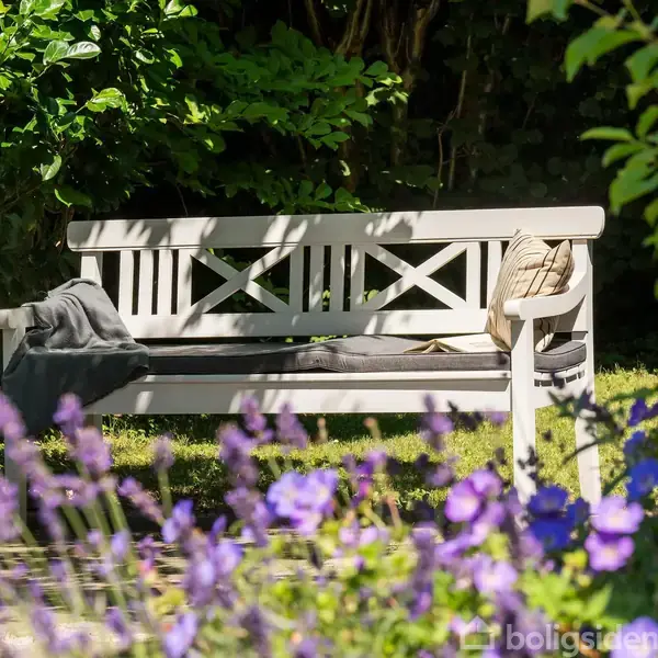 Bænk i træ står i en solrig have omgivet af grønne buske og lilla blomster i forgrunden. En pude og et tæppe ligger på bænken, hvilket skaber hygge.