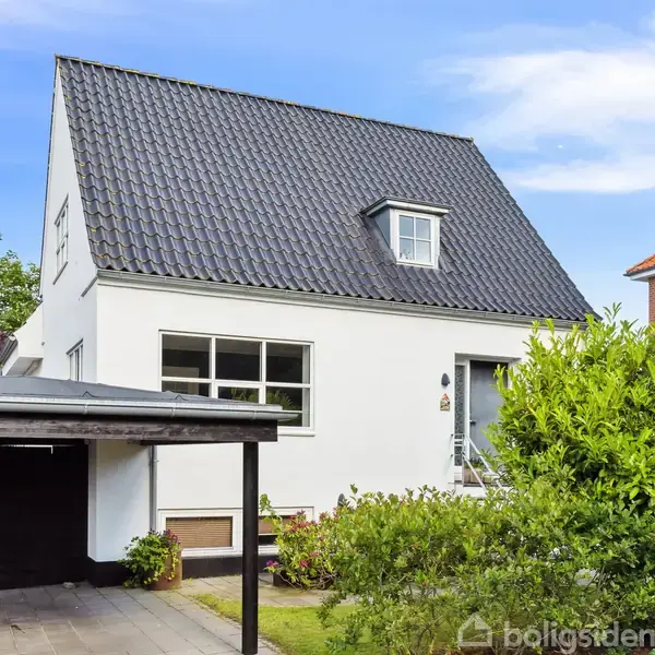 Hvidt hus står stille i en have med grøn busk i forgrunden; sorte tagsten og carport til venstre. Klar blå himmel og nabohus af mursten i baggrunden.