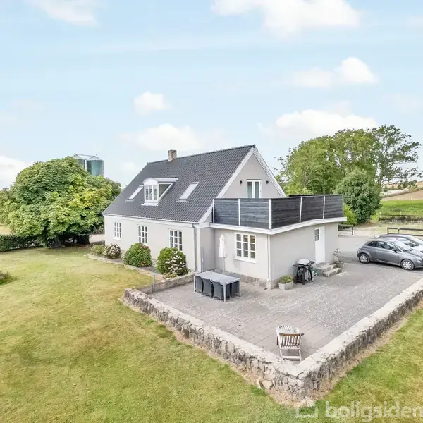 Et hvidt hus med et sort tag står stille i en åben have med grønne buske og træer omkring. To biler er parkeret ved siden af huset.