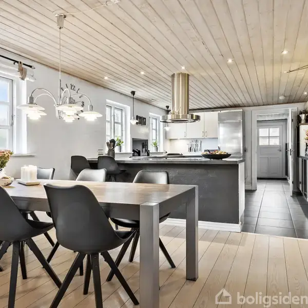 Spisebord i moderne køkken-alrum, omkranset af sorte stole, står på lyse trægulve. Over bordet hænger en pendellampe. Køkkenet har mørke elementer, stålvask og en stor emhætte.