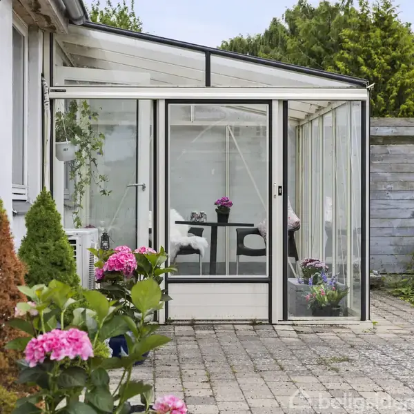 En glasklædt veranda med et bord og blomsterdekorationer befinder sig på en flisebelagt terrasse, omkranset af blomstrende planter og grønne buske.