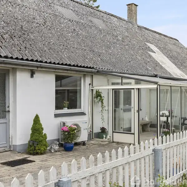 Hus med hvid facade og skråt tag agerer som hovedstruktur, mens en glasindkapslet veranda tilføjer dybde. Foran ses et hvidt stakit og forskellige planter.