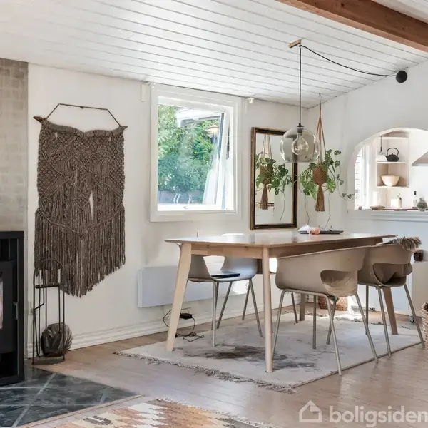 Brændeovn står til venstre, opvarmning. Spisebord med stole midtfor, spisning. Macramé vægdekoration hænger i baggrunden. Træloft og stor kurv ved siden af bordet, hyggelig indretning.
