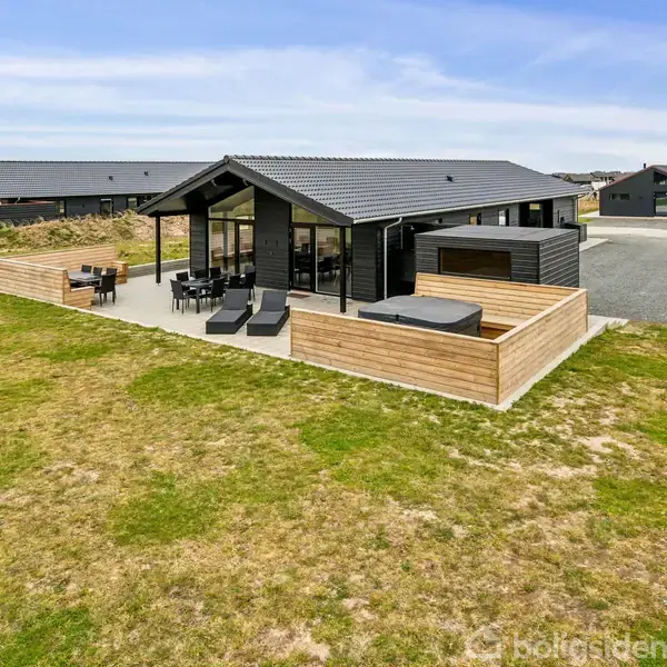 Et sort træhus med store vinduer står på en græsplæne. Foran er en terrasse med udendørs møbler og et jacuzzi. Omkringliggende huse ses i baggrunden.