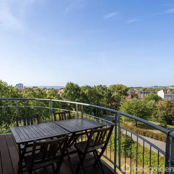 En træterrasse med bord og stole står stille, omgivet af en buet metalrækværk. Udsigten viser en grøn by med huse og spredte træer under en klar blå himmel.