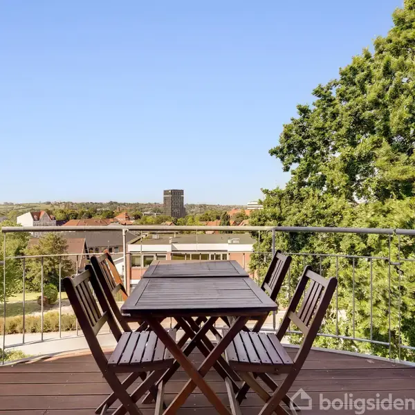 En træterrasse med et bord og fire stole står ovenpå en bygning. I baggrunden ses grønne træer og flere bygninger under en klar blå himmel.