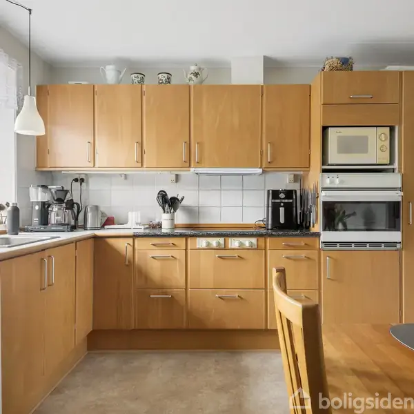 Køkkenskabe i lyst træ monteret på en væg, omkransende en indbygget ovn og mikroovn; bordplade med vask og apparater ved et vindue. Spisebord med stole i forgrunden.