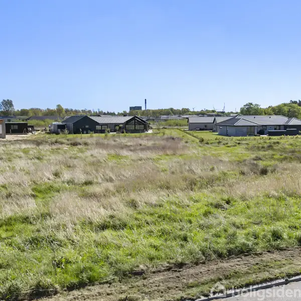 En række lave, moderne huse står stille i et åbent, grønt landskab. Omkring husene strækker græsmarkerne sig ud under en blå himmel, hvilket skaber en fredfyldt atmosfære.