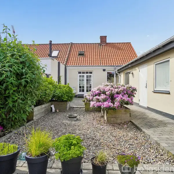 Flere blomster i potter og bed står i en velplejet gårdhave ved et hus med rødt tegltag. Til højre er der en bygning med lyse vægge og hvide døre.