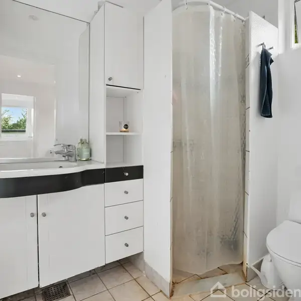 Et hvidt badeværelse med et hjørnebrusebad dækket af et gennemsigtigt forhæng. Ved siden af står et toilet. En vask med et spejl og flere skabe langs væggen.