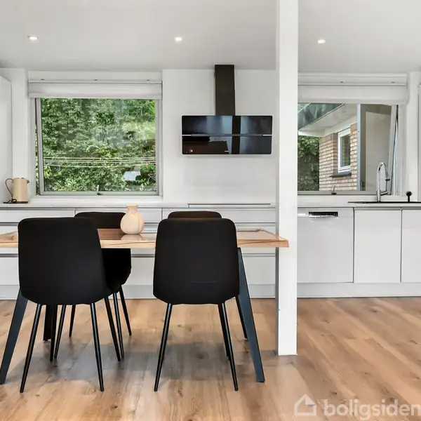 Et moderne, lyst køkken har sorte stole ved et træbord. Køkkenet har hvide skabe, store vinduer med udsigt til naturen og en pejs. Det er rummeligt og elegant indrettet.