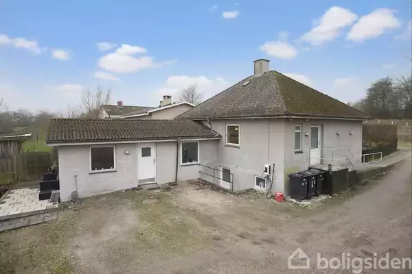 En grå enfamilieshus står stille omgivet af et åbent, grusbelagt område med spredte træer i baggrunden. Huset har et skråt tag og flere vinduer.