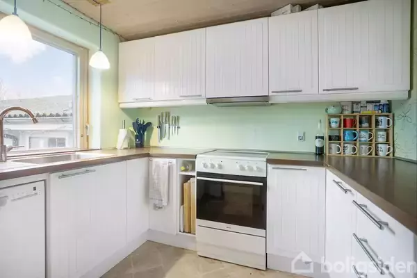 Hvidt køkken står stille, omkranset af skabe med træpaneler og en ovn. Der er kopper på en hylde, og vinduet giver udsigt til et hustag udenfor.