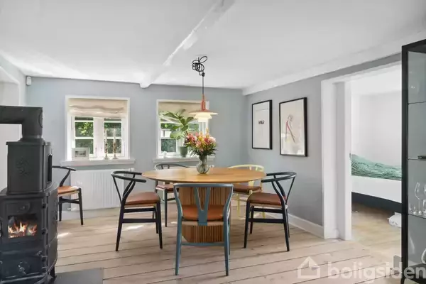 Et rundt spisebord med blomsterbuket i en lys spisestue, omgivet af fire stole. En brændeovn står til venstre, mens vægkunst hænger på de grå vægge.