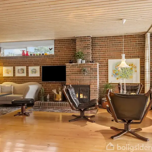 Stue med brune læderstole og sofa, arrangeret omkring et fjernsyn og en pejs. Væggene er af mursten, og der er store vinduer, som giver rummet naturligt lys. Planter og malerier dekorerer rummet.