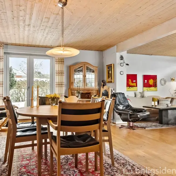 En spisestue indeholder et rundt træbord med stole, placeret på et orientalsk tæppe. Ved siden af står en glasvitrine, mens en sofa og lænestole ses i baggrunden.