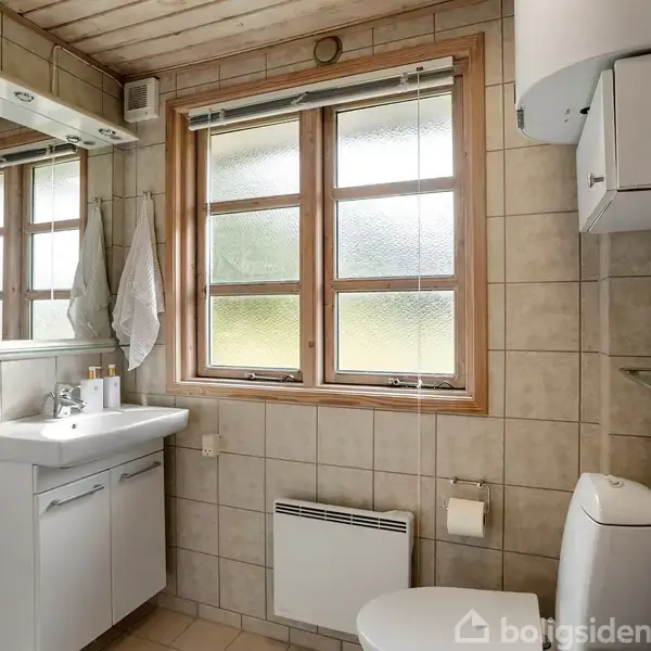 Badeværelse med vask og spejl på venstre side, toilet til højre. Flisebelagt væg og gulv, vindue i midten giver naturligt lys. Hylder og radiator under vinduet.