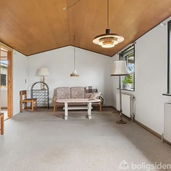 Stue med sofa og lille bord, lampen hænger fra loftet. Store vinduer med udsigt til verandaen. Gulvlampe ved siden af sofaen, og fladskærm på væggen.