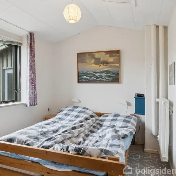 Seng står i midten af et lyst værelse med et mønstrede tæppe og plaid dyner. Et vindue og maleri med havtema ses på væggen. En lampe hænger fra loftet.