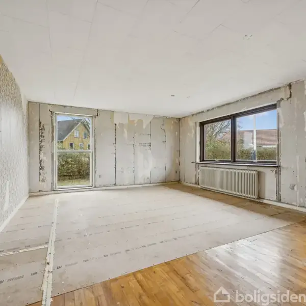 Værelse med afskallet tapet og træ- og beton/linoleumgulv. Store vinduer på to vægge viser en udendørs have og hus. Rummet virker klart til renovering. Ingen tekst bortset fra "Boligsiden."