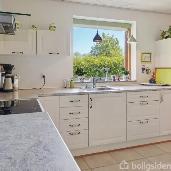 Køkkenbord med hvidt skab står stille under et åbent vindue. Udenfor ses grønt løv. På bordet er en kaffemaskine og opvaskemiddel, mens et bordhjørne med en dug ses til højre.