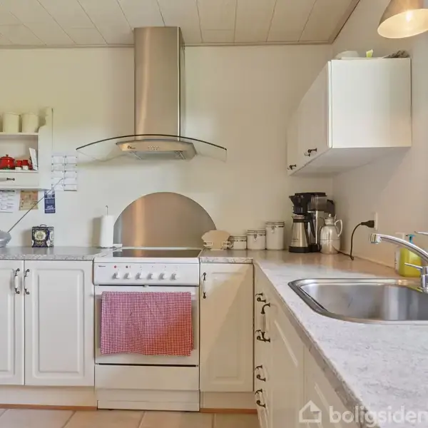 Et køkken med en hvid komfur og emhætte, omgivet af hvide skabe og en vask. En plante og kaffemaskine står ved vinduet med lysindfald. Hylder med dekorative genstande.