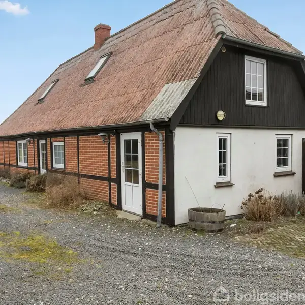 Et rødt murstenshus med stråtækt tag står på en grusbelagt grund. Det har hvide døre og vinduer. Omgivet af marker under en blå himmel.
