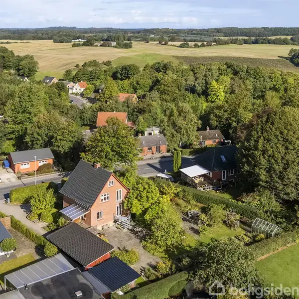 Et rødt murstenshus står midt i en landsby med frodige grønne træer rundt om. Omgivet af naboer og landbrugsmarker i baggrunden. Skyfri himmel ovenover.