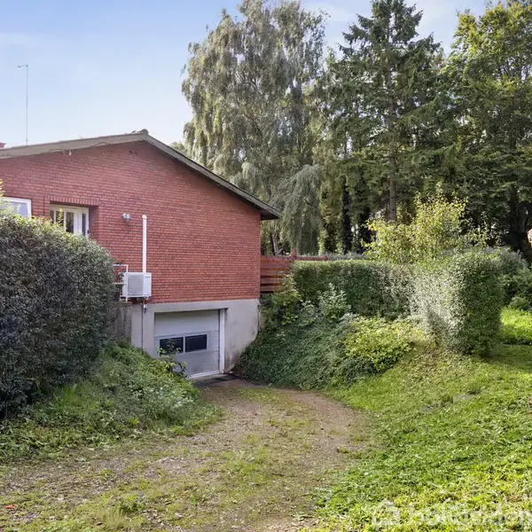 Rødt murstenshus med en hævet indgang. En grusvej fører til en garage i kælderniveau. Haven er omkranset af buske, med en udskåret træstub som dekoration nær vejen.
