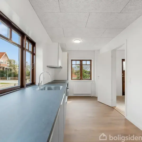 Køkken med lyse overflader og moderne design, står stille. Store vinduer giver udsigt til et rødt taghus udenfor. Rummet er lyst og rummeligt med trægulv.