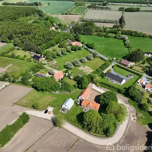 Flere rødtage bygninger ligger blandt frodige grønne marker og træer. Grusveje forbinder ejendommene i det landlige område.