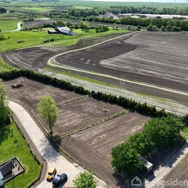 Markeret område med pløjet jord, omgivet af grøn beplantning og en grusvej i nærheden. En stor pløjemark strækker sig i baggrunden med landevej og bygninger i det fjerne.