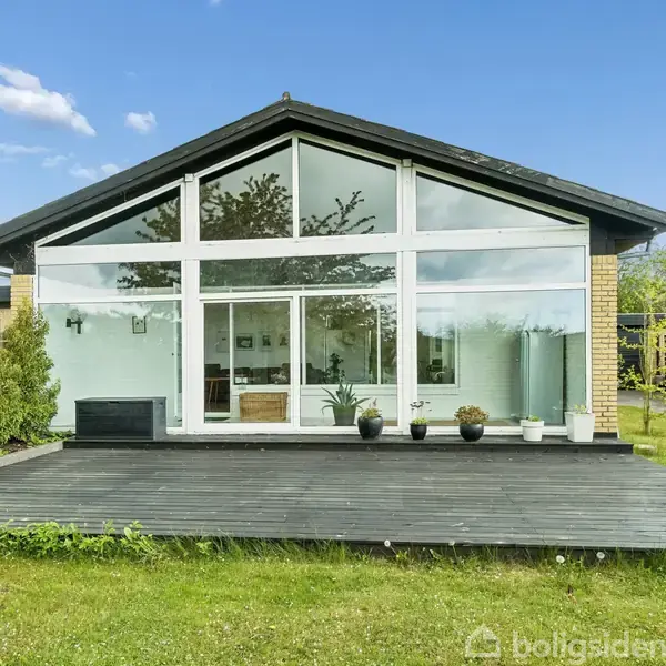 Et moderne hus med store glaspartier står på en terrasse omgivet af grønne græsarealer og buske. Himlen er klar med enkelte skyer.