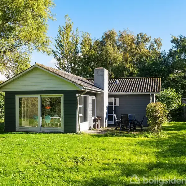 Sommerhus med store vinduer står på en grøn græsplæne omgivet af træer. Udendørs terrasse med havemøbler og grill ses til højre.